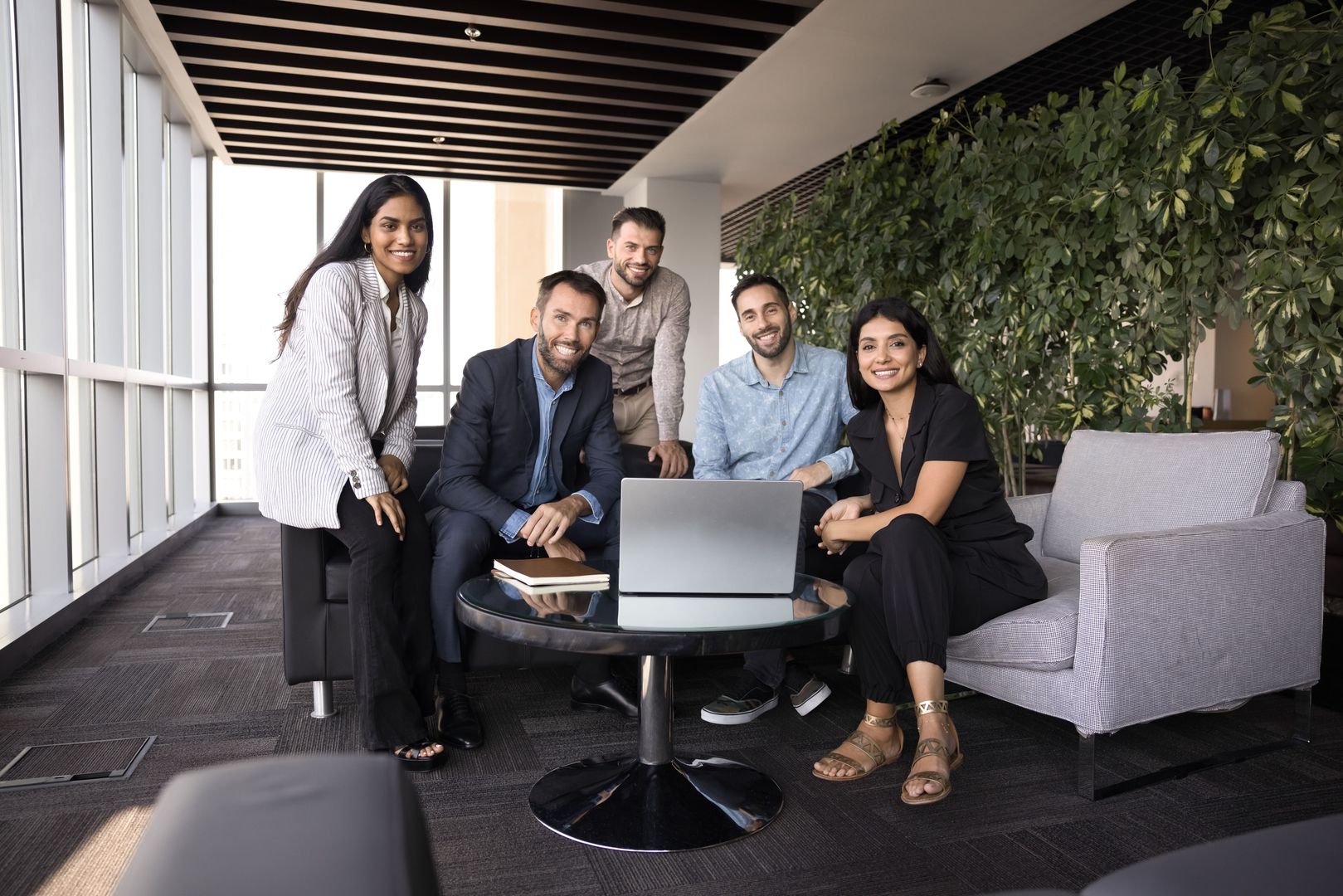 Fünf Personen sitzen gemeinsam in einem modernen Bürobereich um einen Tisch mit Laptop.